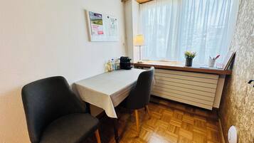 A small, tidy room with a table and two chairs, a window with sheer curtains, a wall-mounted lamp, and a radiator.