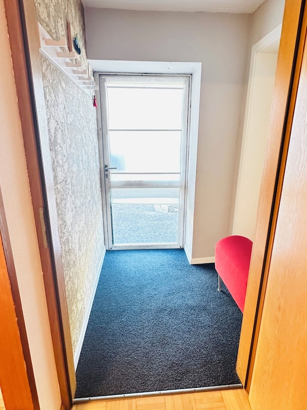 A narrow hallway with a wooden floor, a red chair, and a door leading outside.