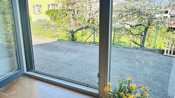 Ein Fenster mit Blick auf ein gelbes Gebäude, ein Blumentopf mit gelben Blüten und ein klarer, blauer Himmel.