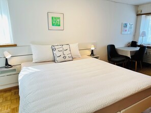 Habitación de hotel con una cama, una mesita de noche con lámpara, una silla, un escritorio con lámpara y un cuadro en la pared.