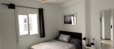 A bedroom with a bed, a ceiling fan, a window with curtains, a bedside table, and a wall-mounted mirror.