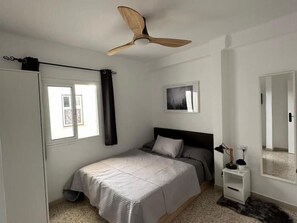Ein Schlafzimmer mit Bett, Deckenventilator, Fenster mit Vorhängen, Nachttisch und wandmontiertem Spiegel.