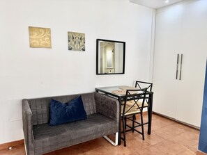 Um quarto pequeno com um sofá cinzento, uma mesa com cadeiras, um espelho e dois armários fixados na parede.