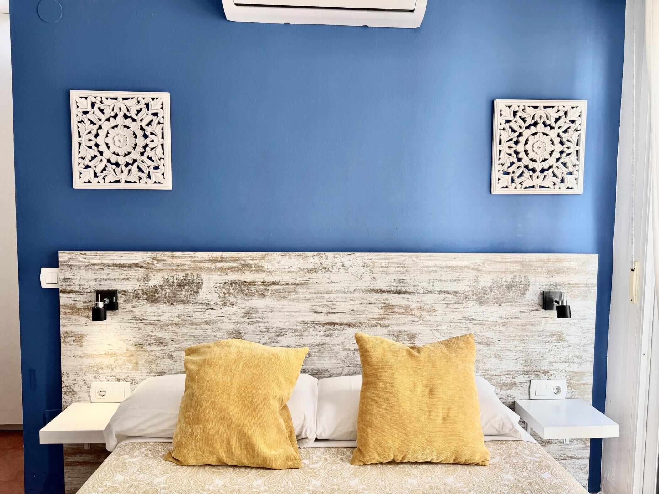 Una habitación con pared azul, dos paneles decorativos, cabecera de madera y dos almohadas amarillas sobre una cama blanca.