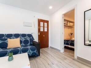 Ein Zimmer mit einer Couch, einem kleinen Tisch mit einer Pflanze, Holzboden, einer Tür und einem Schrank mit Regalen.