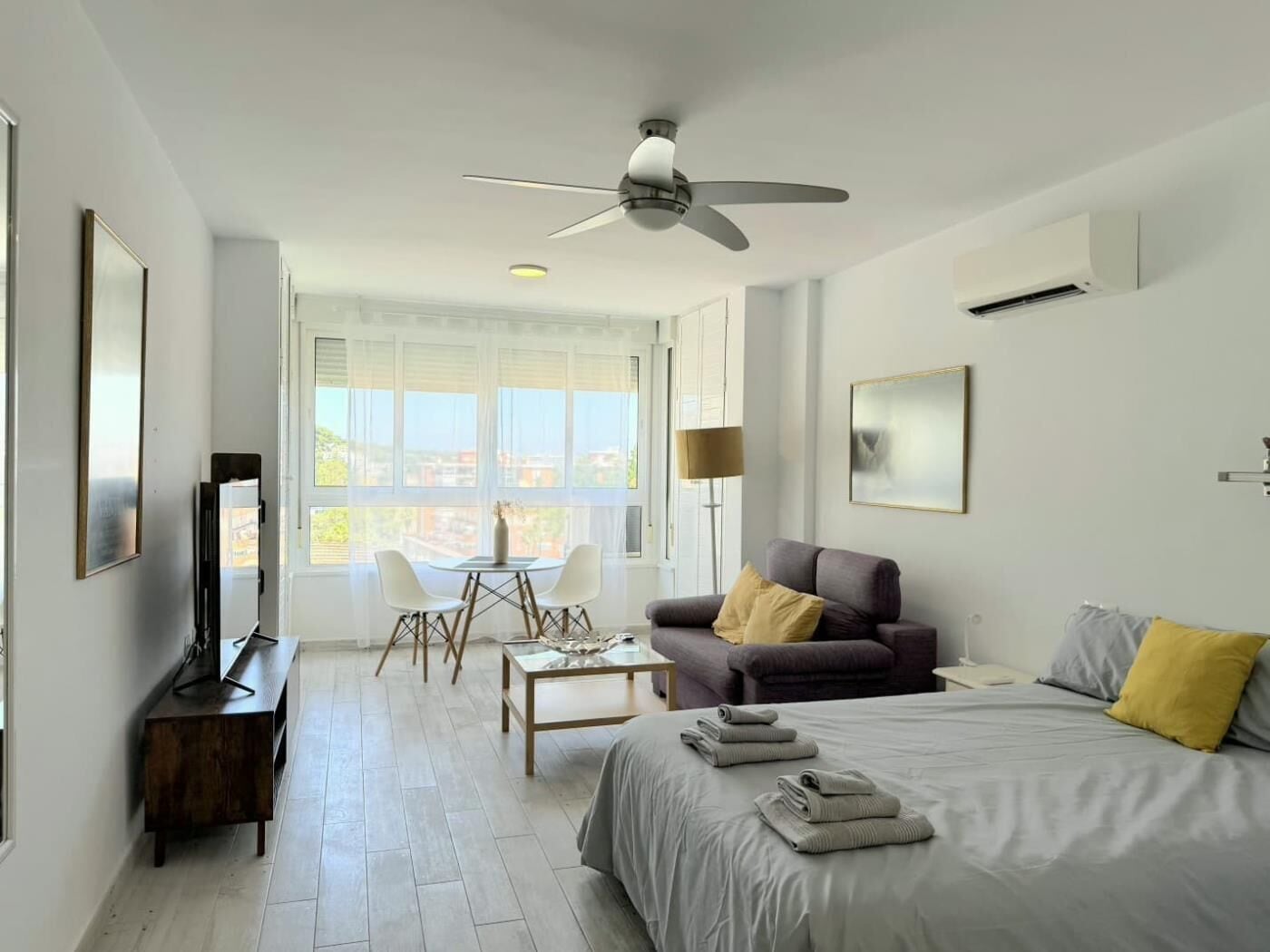 A modern hotel room with a bed, a sofa, a dining table with chairs, a ceiling fan, and a large window.