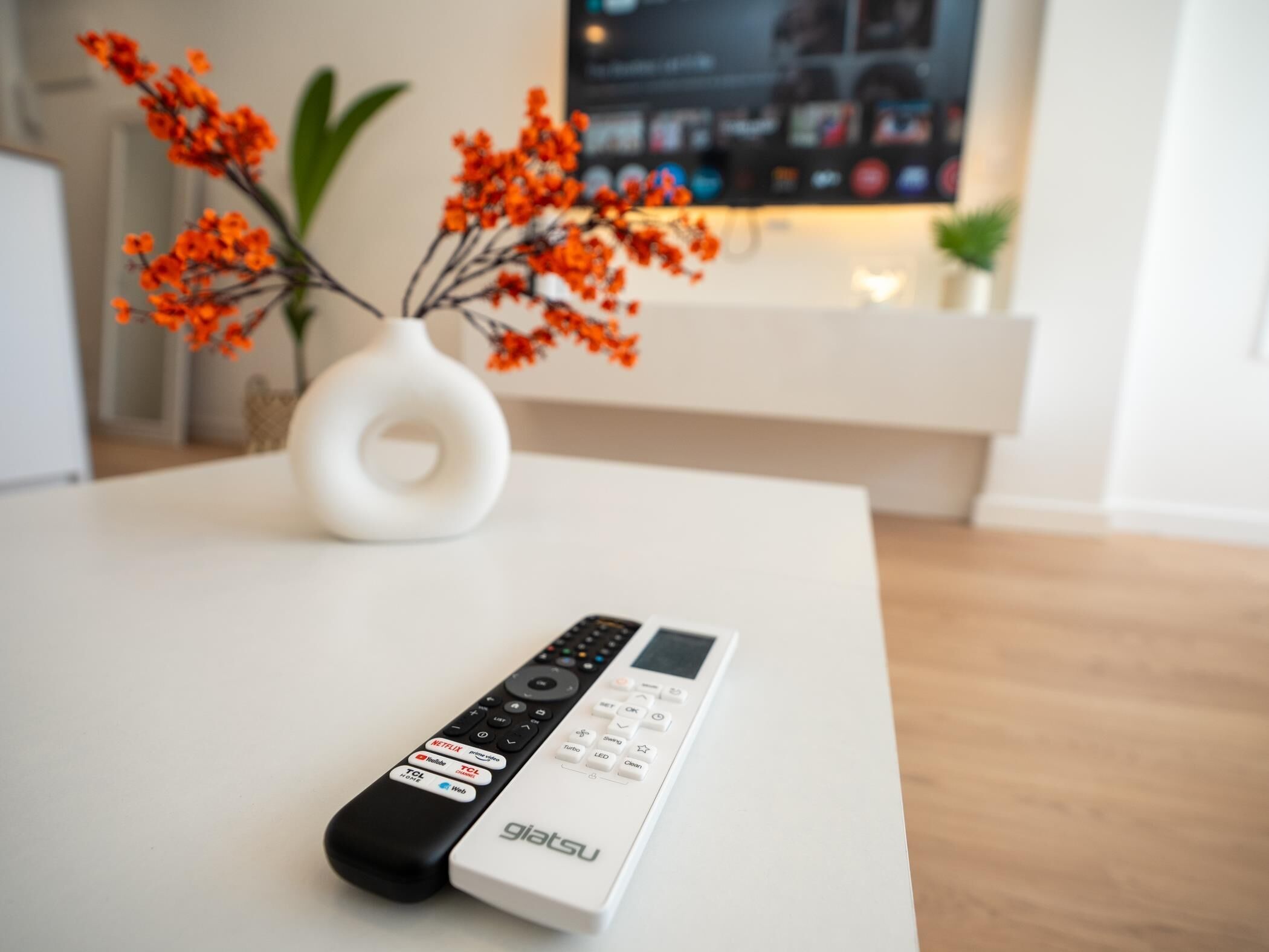 A remote control on a white table with a vase of orange flowers and a flat-screen TV in the background.