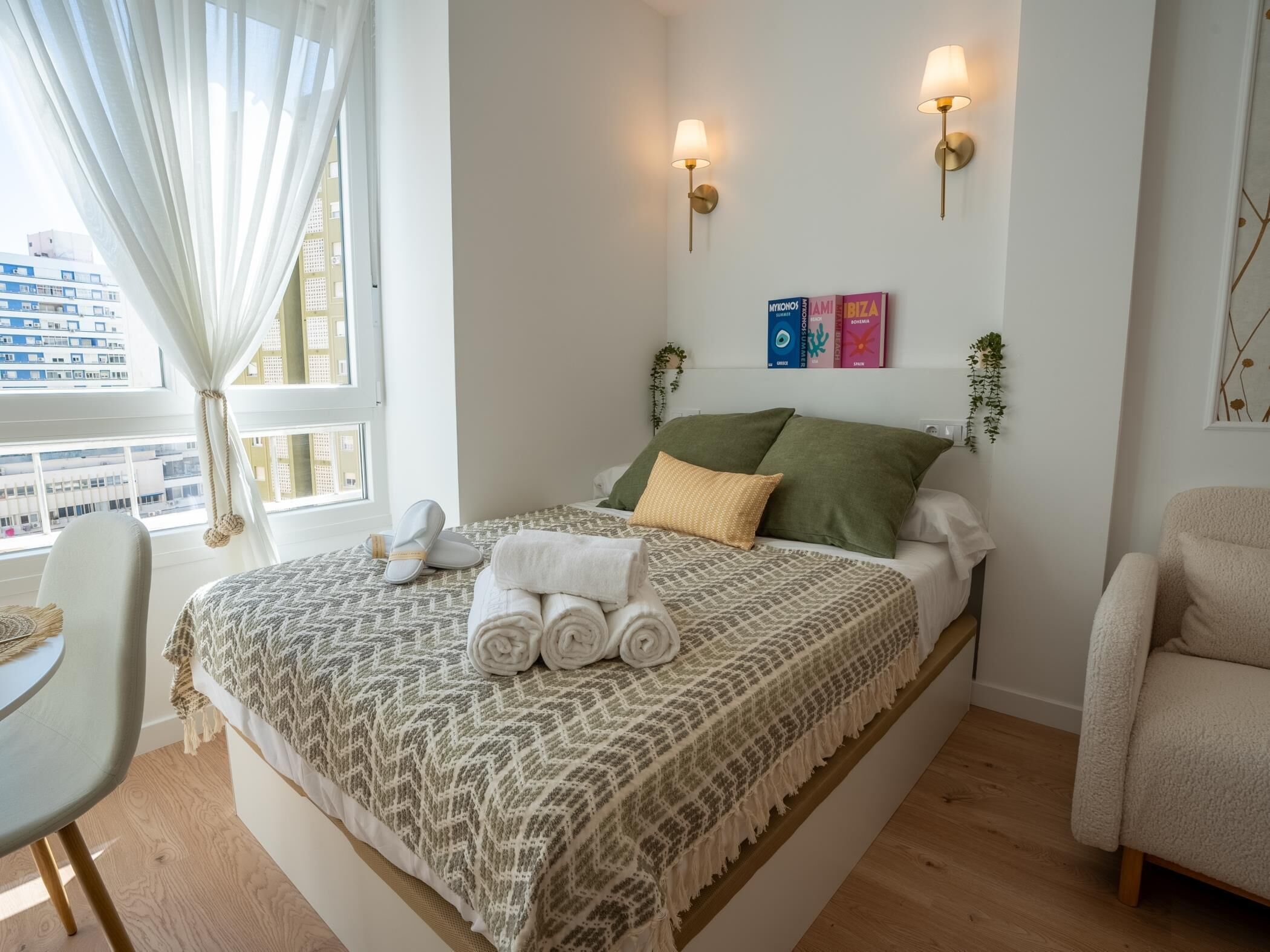 Habitació d'hotel amb llit, cadira, taula de nit, finestra amb cortines i làmpada penjada a la paret.