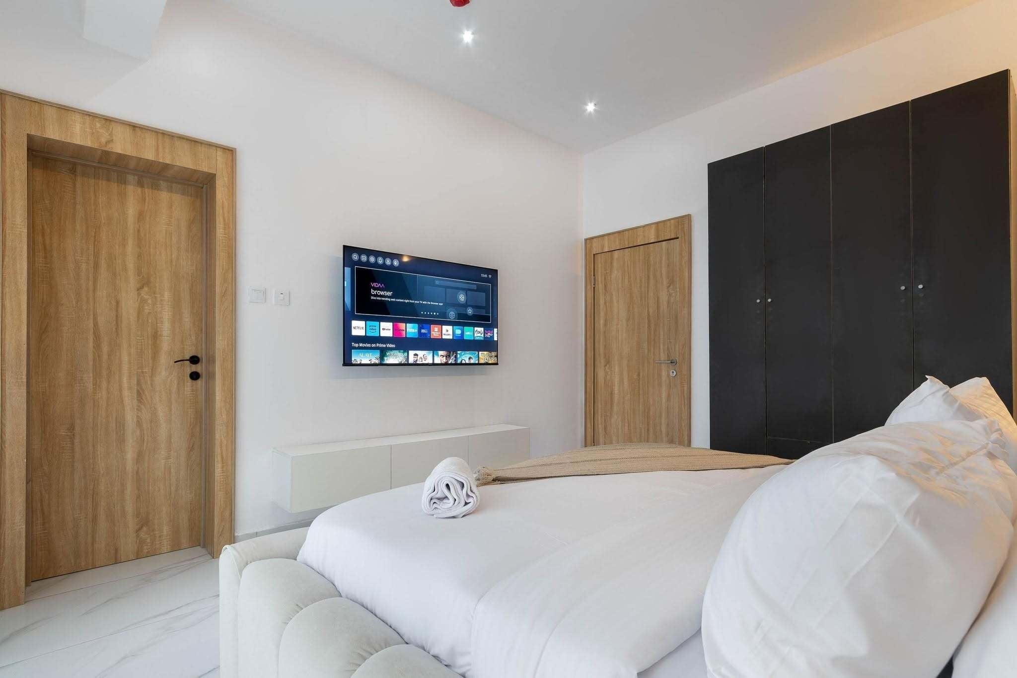 A modern bedroom with a large bed, a mounted TV displaying a streaming service interface, and a wooden wardrobe.