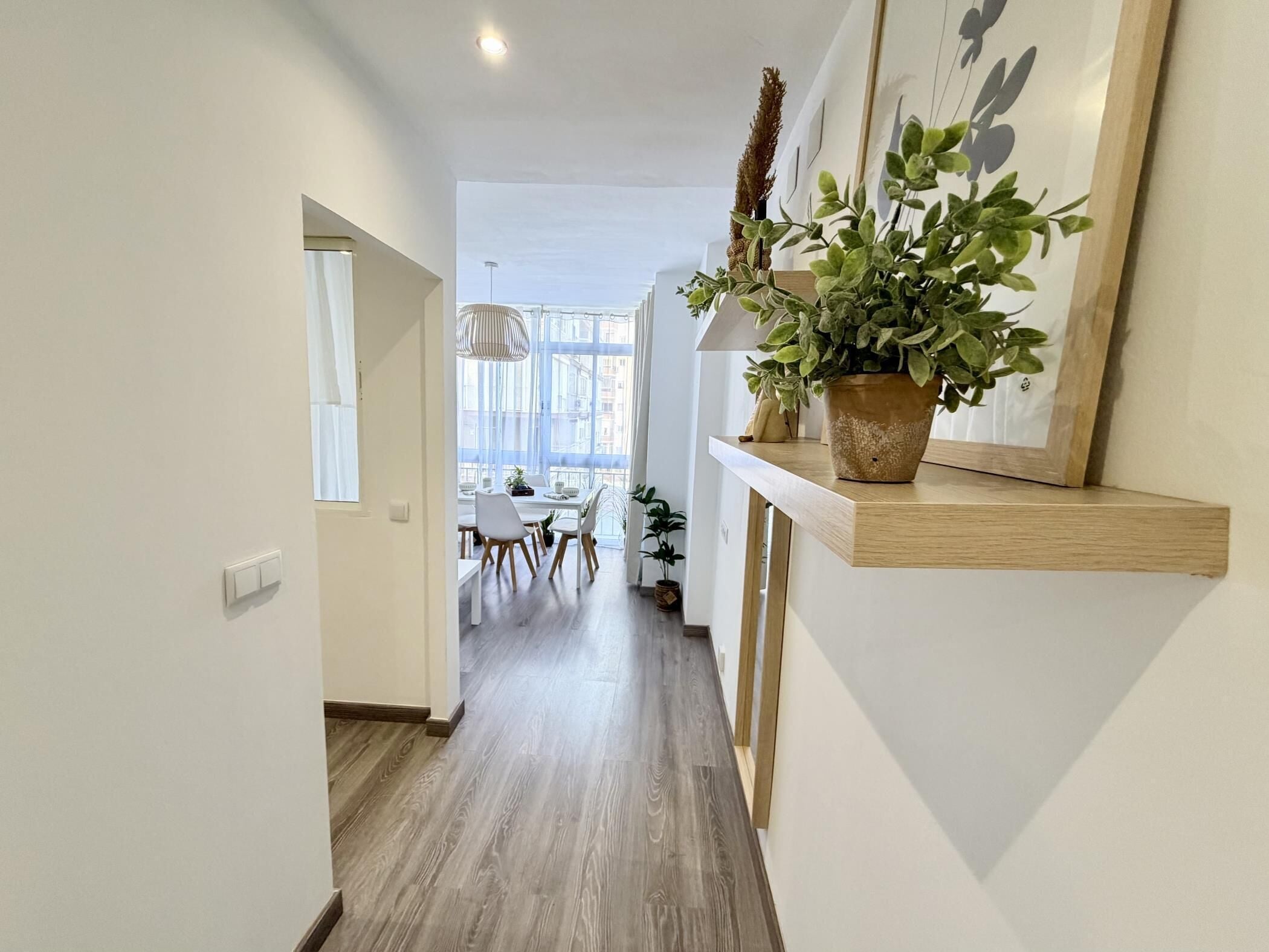 Un pasillo con suelo de madera, un comedor al fondo y una planta en un estante.