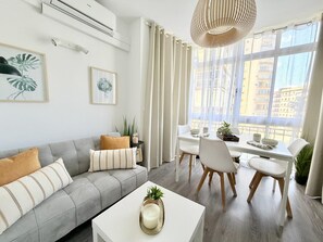 Een moderne woonkamer met een grijze bank, een witte salontafel en een eethoek met een witte tafel en stoelen. Er hangen twee kunstwerken aan de muur, een plafondlamp en een raam met gordijnen.