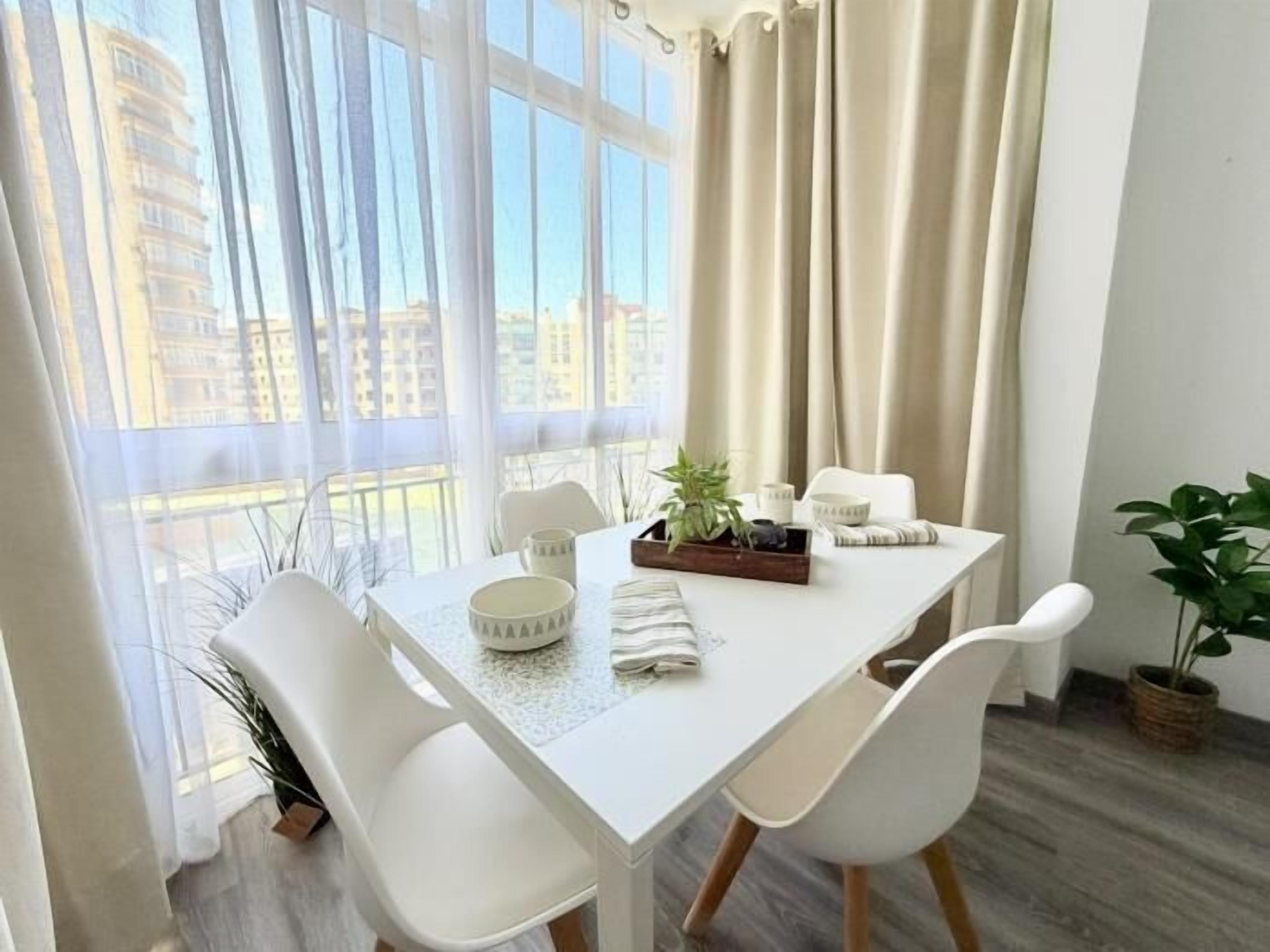 Un moderno comedor con una mesa y sillas blancas, una bandeja con plantas y una vela, y vistas a edificios a través de grandes ventanales.