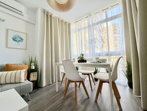 Een moderne woonkamer met een eethoek, met een bank, stoelen, een tafel en een groot raam met gordijnen.