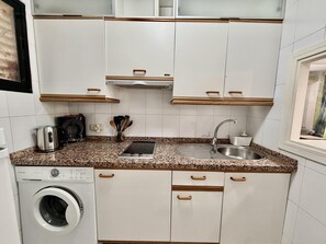 Cocina compacta con armarios blancos, lavadora integrada, un lavamanos y una cafetera.