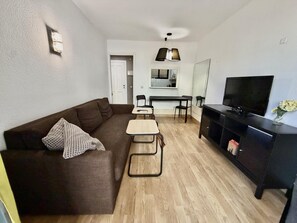 A compact living space with a sofa, a small dining table, a TV, and a kitchenette in the background. - Ground-Floor Apartment with Private Terrace 125 m from the Beach, La Colina (Torremolinos)