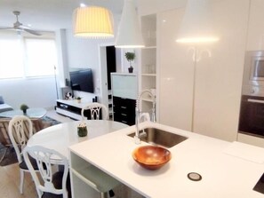 Een moderne keuken met een wit aanrecht, een roestvrijstalen spoelbak en een kom. Er is een eethoek met witte stoelen en een tafel.