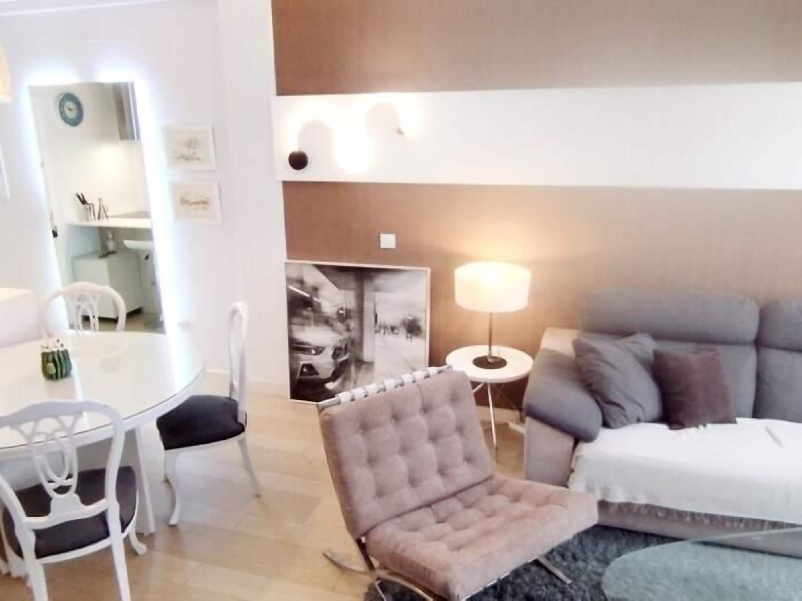 Ein modernes Wohnzimmer mit einem grauen Sofa, einem gesteppten Sessel, einem runden Couchtisch und einem Beistelltisch mit Lampe.