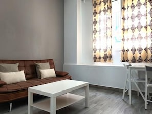 A living room with a brown sofa, a white coffee table, and a window with patterned curtains. - Brand-New Studio 130 m from Bajondillo Beach, Torremolinos (Torremolinos)