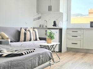 Een knus leefruimte met een bed, een klein tafeltje, een plant en een kitchenette.