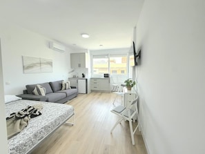Een moderne, goed verlichte kamer met een bed, een grijze bank, een kleine kitchenette en een eethoek met een witte stoel en tafel.