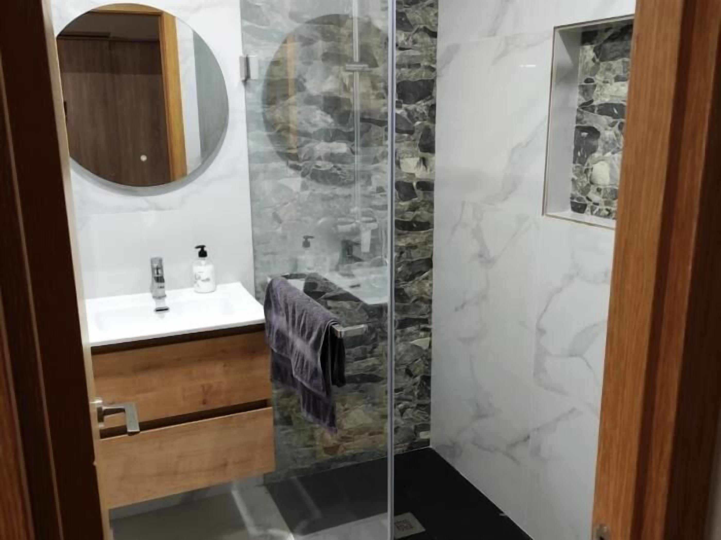 A modern bathroom with a glass shower enclosure, a wooden vanity, and a marble wall.