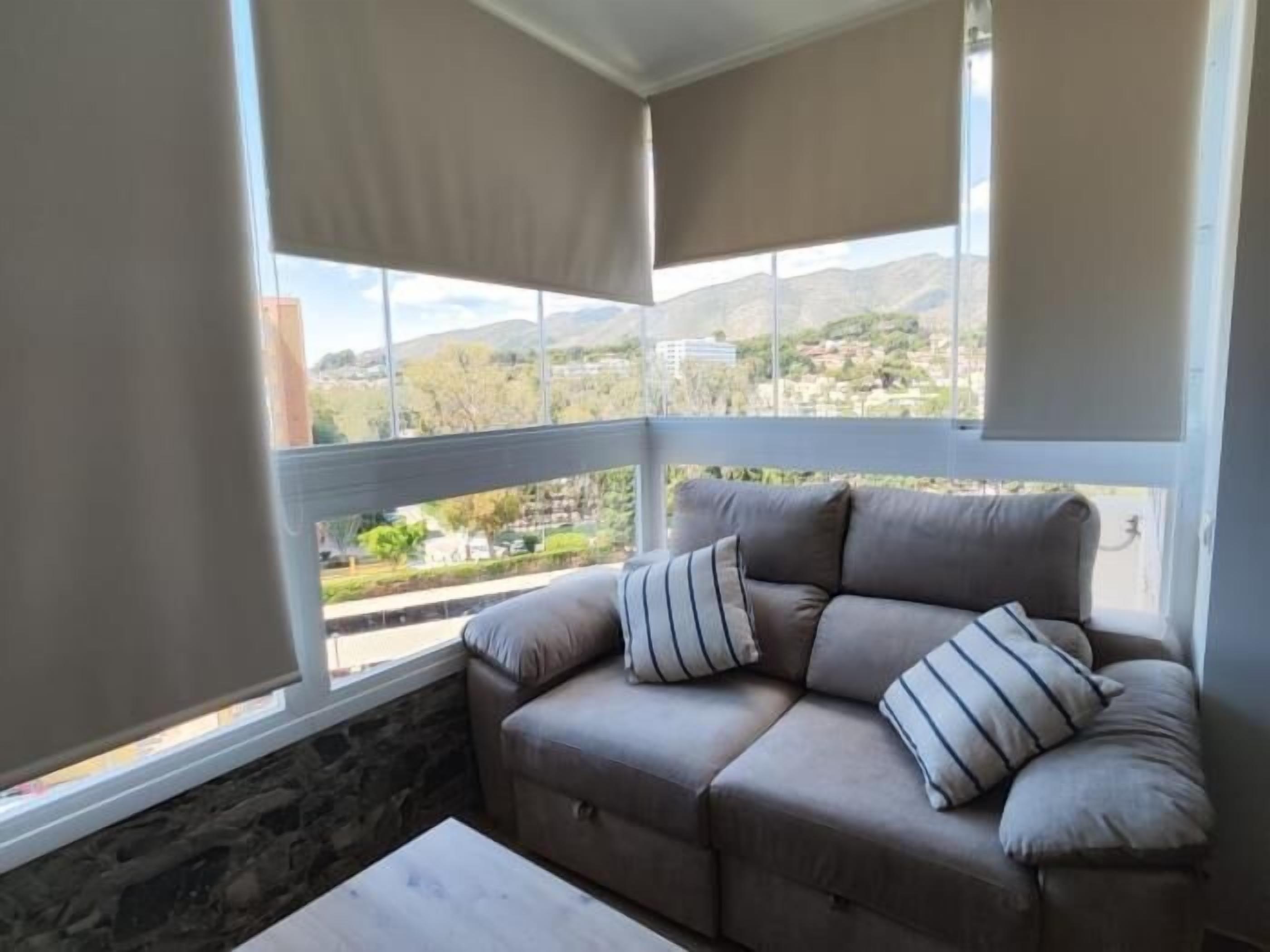 Une pièce d’angle avec un canapé beige, deux coussins à rayures et une vue sur la ville et les montagnes, grâce à de grandes fenêtres.