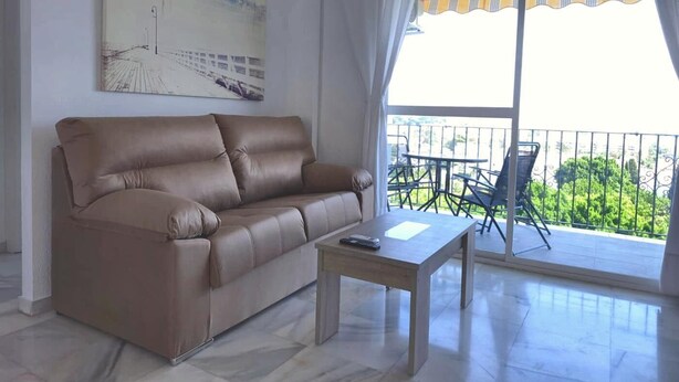 Sala de estar com um sofá marrom, uma mesinha e vista para uma varanda com mobiliário de exterior.