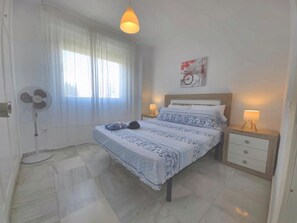 Ein Schlafzimmer mit einem Bett, Nachttischlampen, einem Ventilator, einem Fenster mit Vorhängen und einem Wandbild.