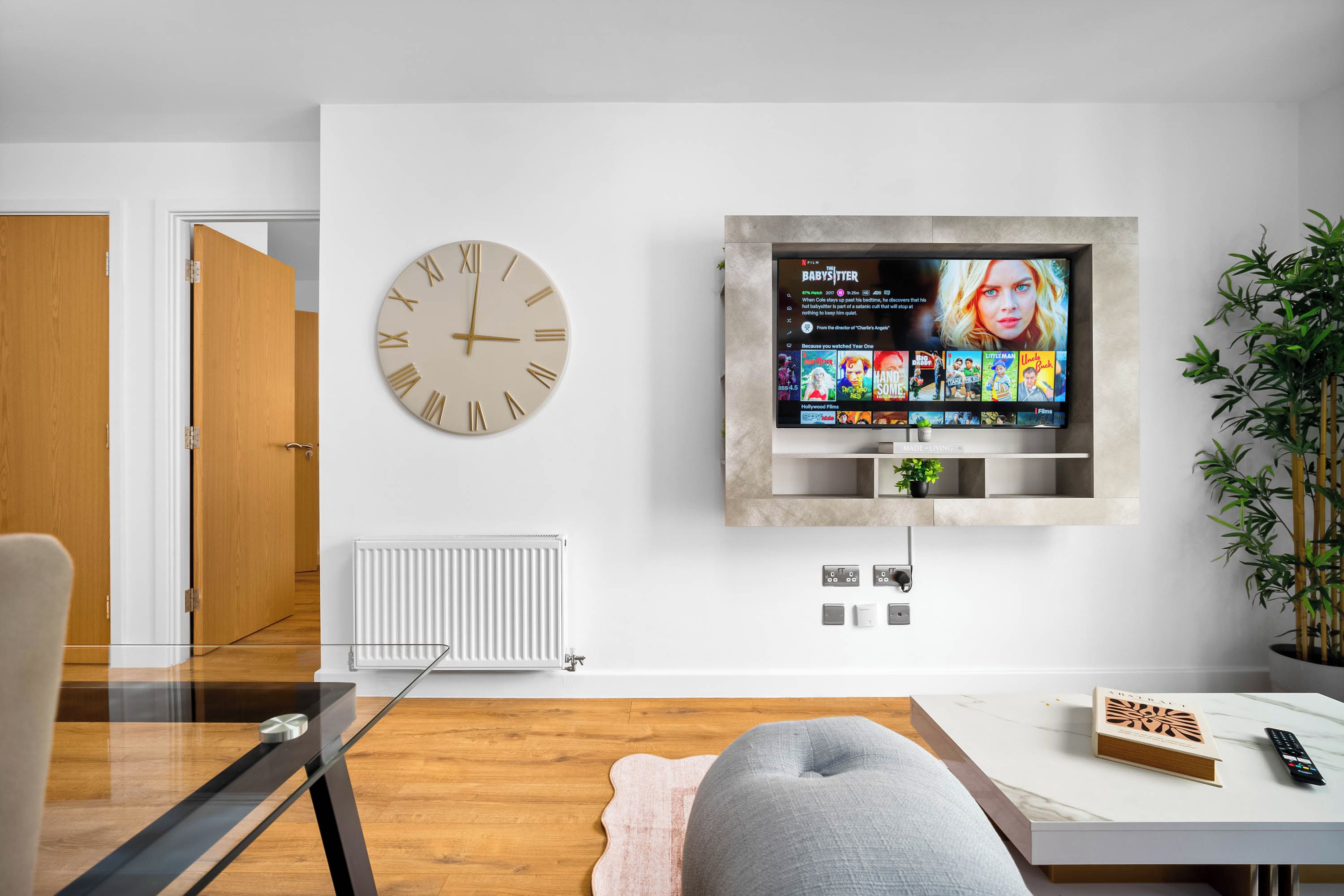 Un salón moderno con un televisor de pantalla montado en la pared mostrando una selección de películas, un reloj redondo en la pared y un radiador.