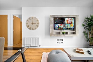 Ein modernes Wohnzimmer mit an der Wand montiertem Fernseher, der eine Filmsammlung zeigt, einer runden Wanduhr und einer Heizung.