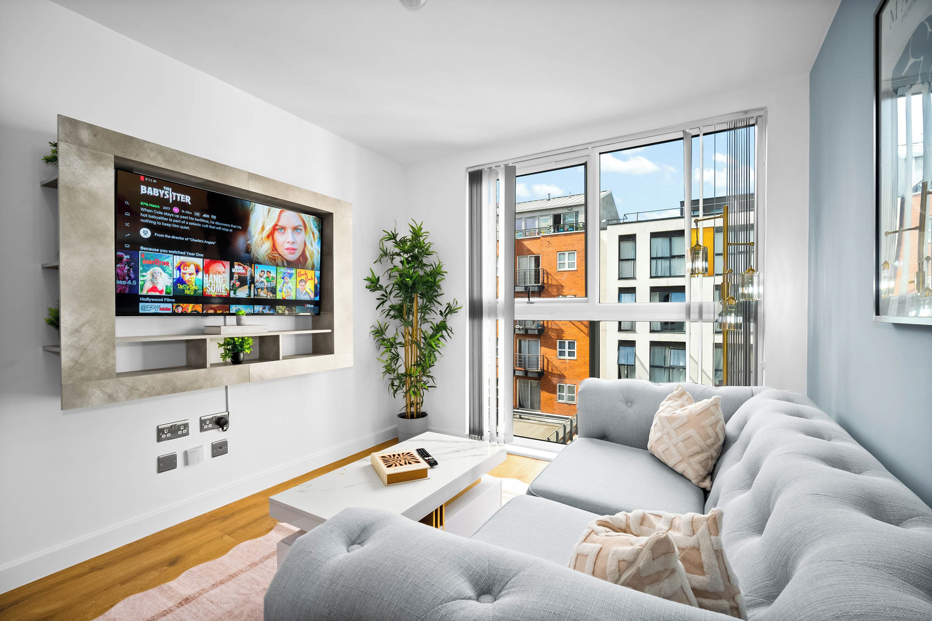 A modern living room with a large flat-screen TV mounted on the wall, a grey sofa, a small coffee table, and a potted plant.