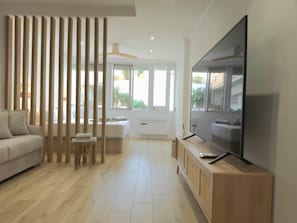 A modern living room with a sofa, a TV on a wooden console, a large window, and a ceiling fan. - Renovated Studio with Pool and Terrace, 7 Minutes from Bajondillo Beach (Torremolinos)