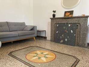 A living room with a grey sofa, a small side table, a fireplace with a decorative mantle, and a patterned rug. - One of a kind! In a Provençal village, a spacious apartment with access to our bookstore (Courthézon)