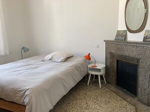 A bedroom with a bed, a small table with a lamp, a fireplace, and a mirror on the wall. - One of a kind! In a Provençal village, a spacious apartment with access to our bookstore (Courthézon)