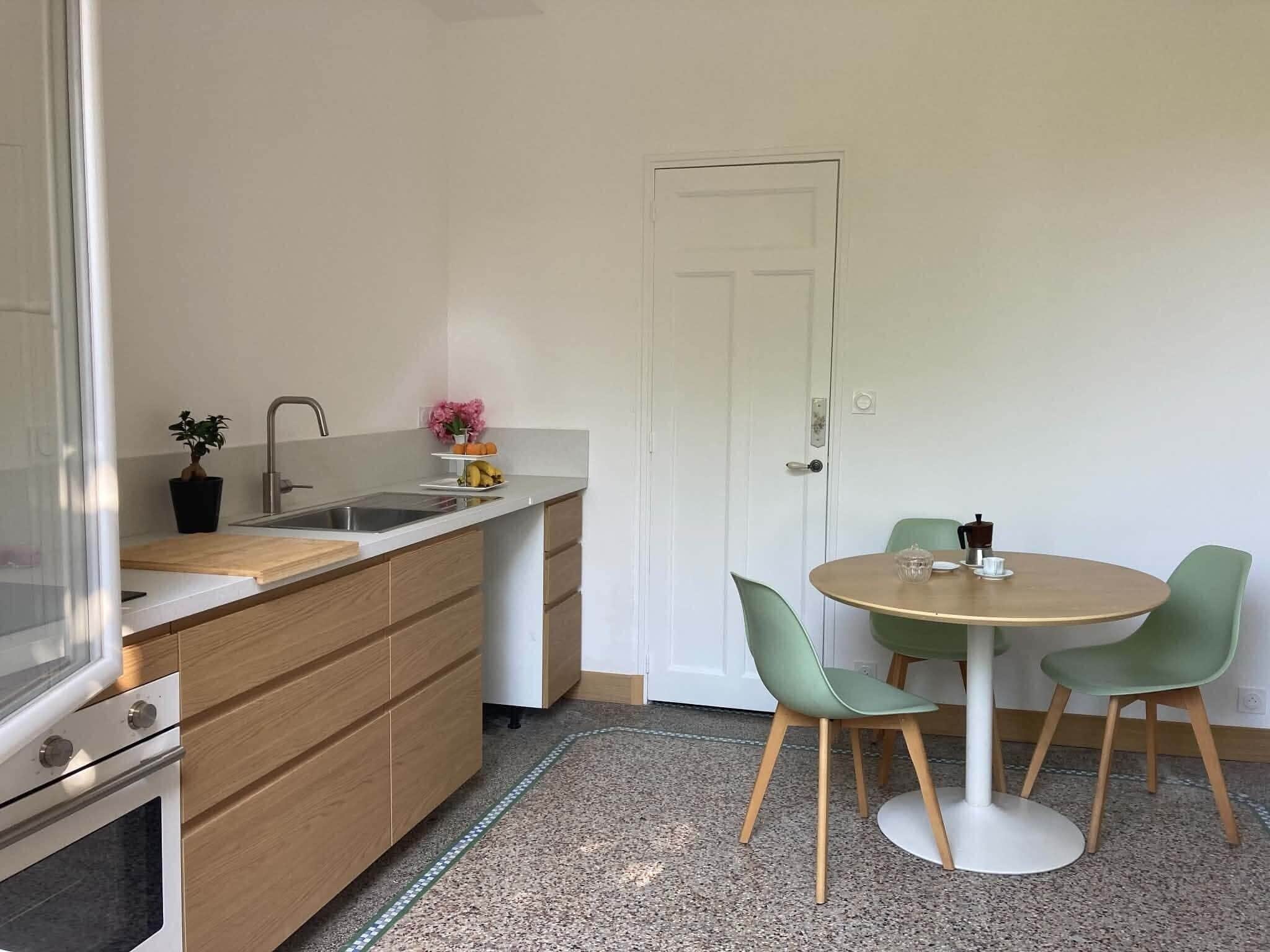 Eine kleine Küche mit Spüle, Arbeitsfläche und Holzschränken. Es gibt einen Essbereich mit einem runden Tisch und zwei Stühlen.