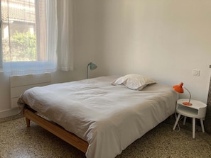 A simple, unadorned bedroom with a bed, a nightstand, a lamp, and a window with sheer curtains. - In a Provençal village, a spacious apartment with access to our bookstore (Courthézon)