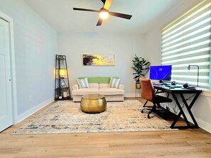 Ein Wohnzimmer mit einer Couch, einem Couchtisch, einem Schreibtisch mit Computer sowie einem Deckenventilator und einem Fenster mit Jalousien.