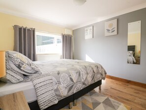 Ein Schlafzimmer mit Bett, Nachttisch, Fenster mit Vorhängen, Wand mit Kunstwerk und Holzboden.
