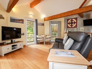A living room with a flat-screen TV, a leather sofa, a dining table with a magazine, and a balcony with a view of the sea. - Brambleside Lodge (Truro)