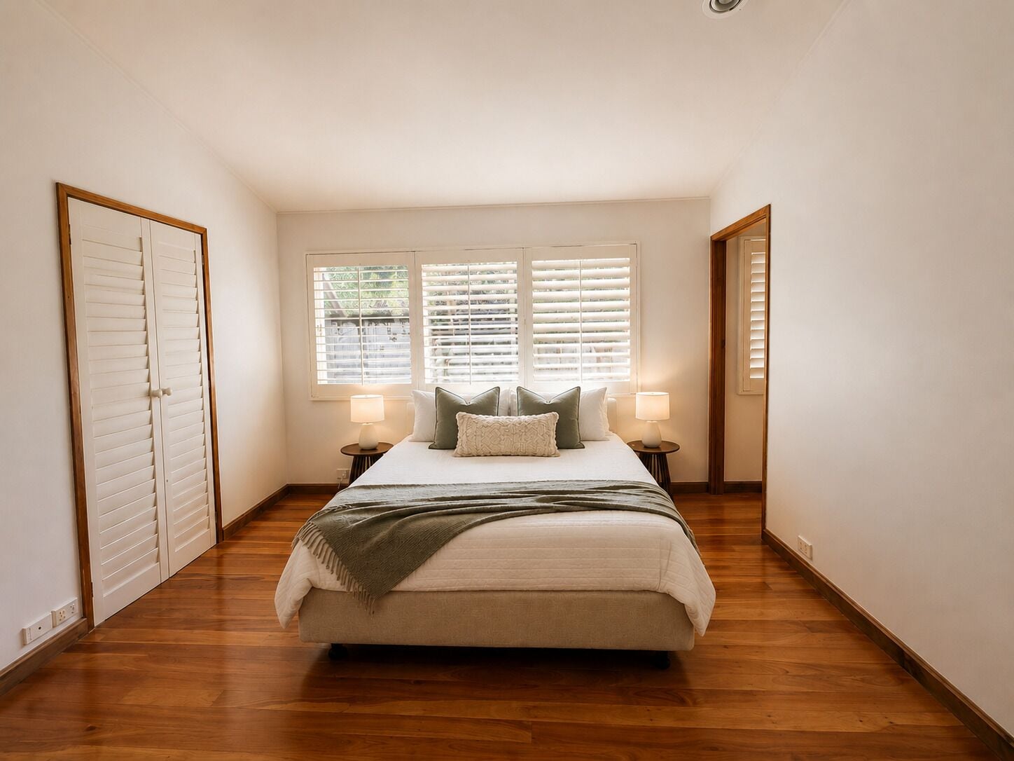 A bedroom with a bed, two bedside lamps, a window with blinds, and a door.