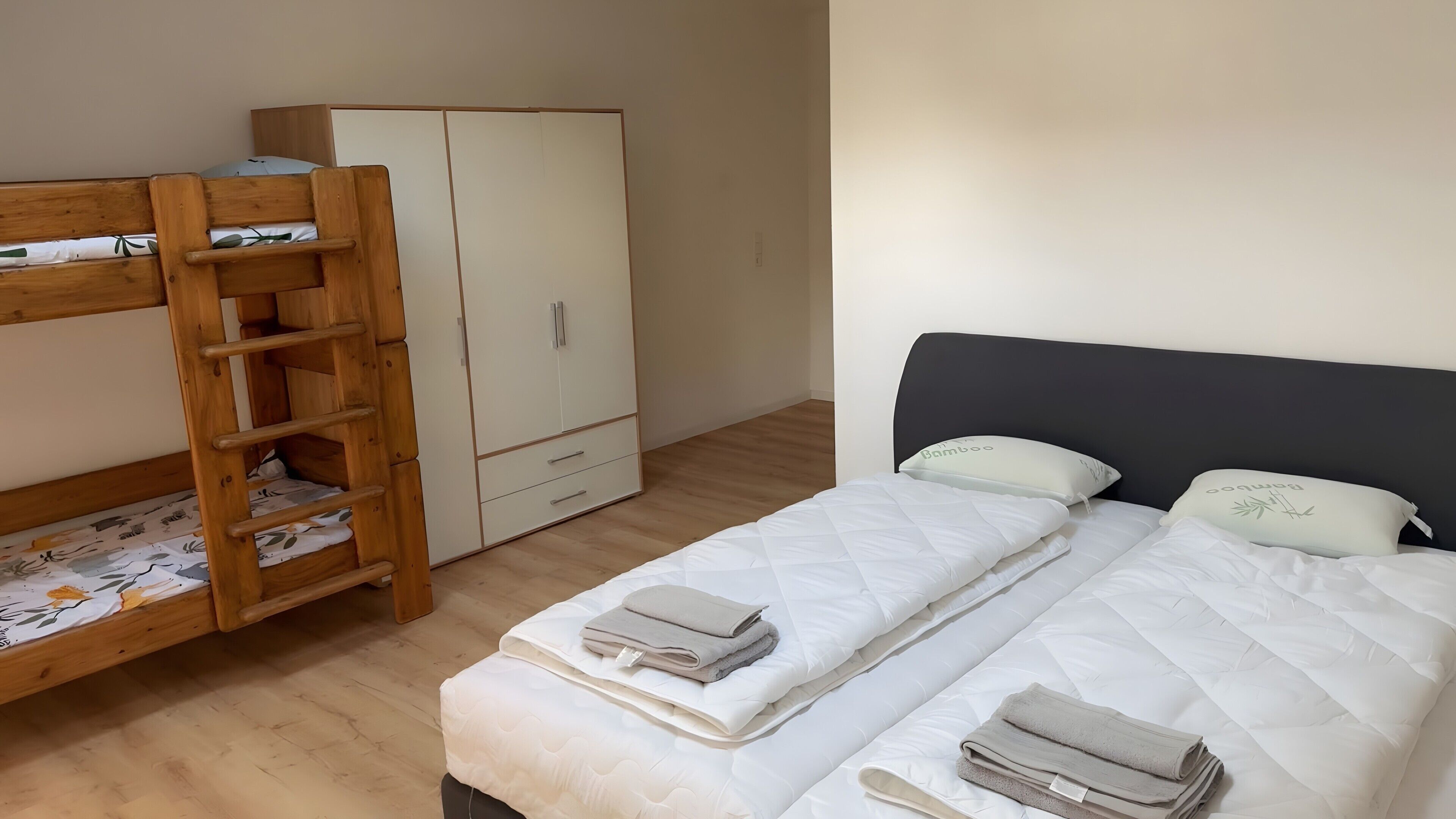 A bedroom with a bunk bed, a wardrobe, and a bed with pillows and folded towels.