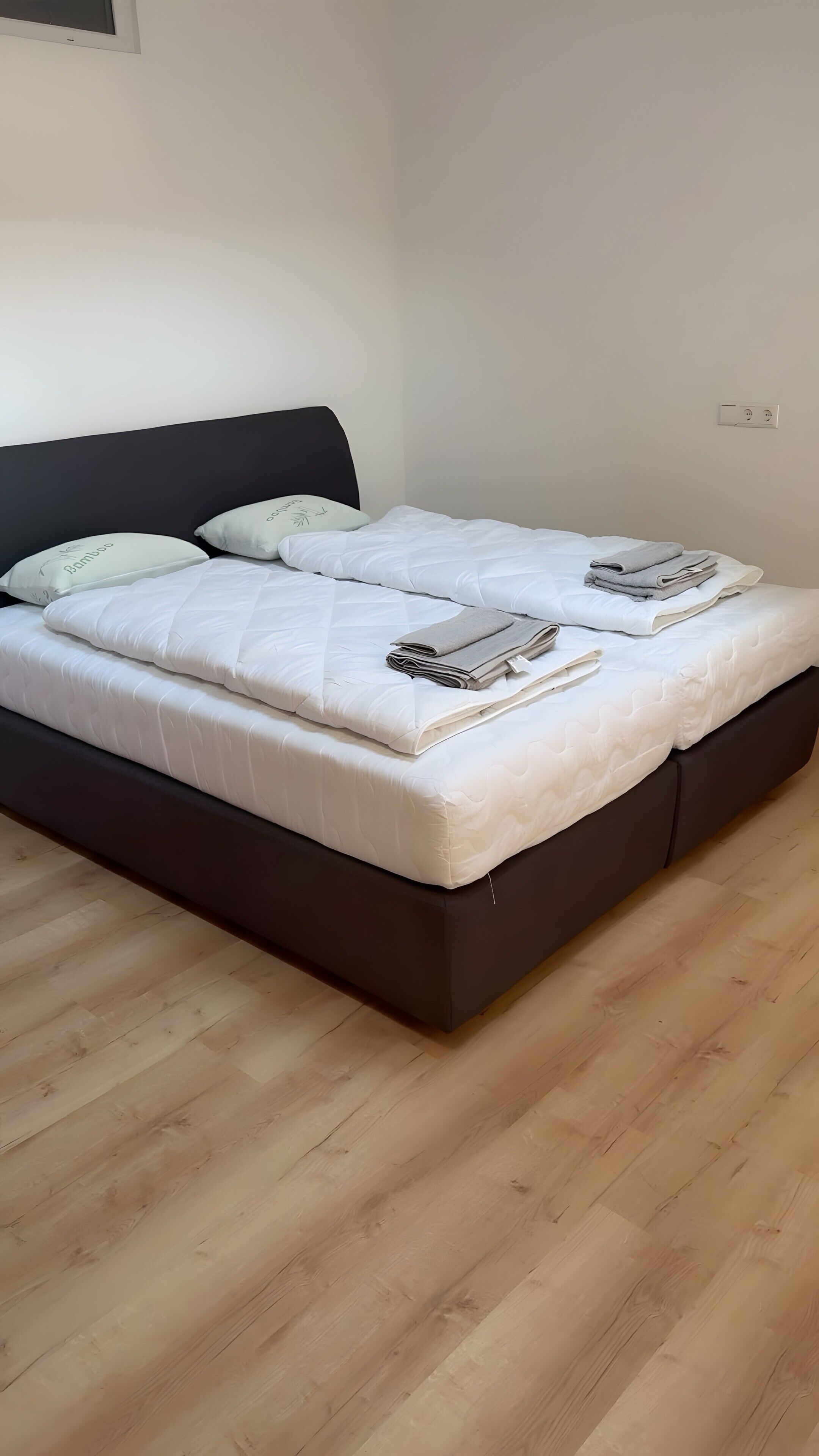 A bed with white bedding and two pillows, a dark headboard, and a wooden floor.