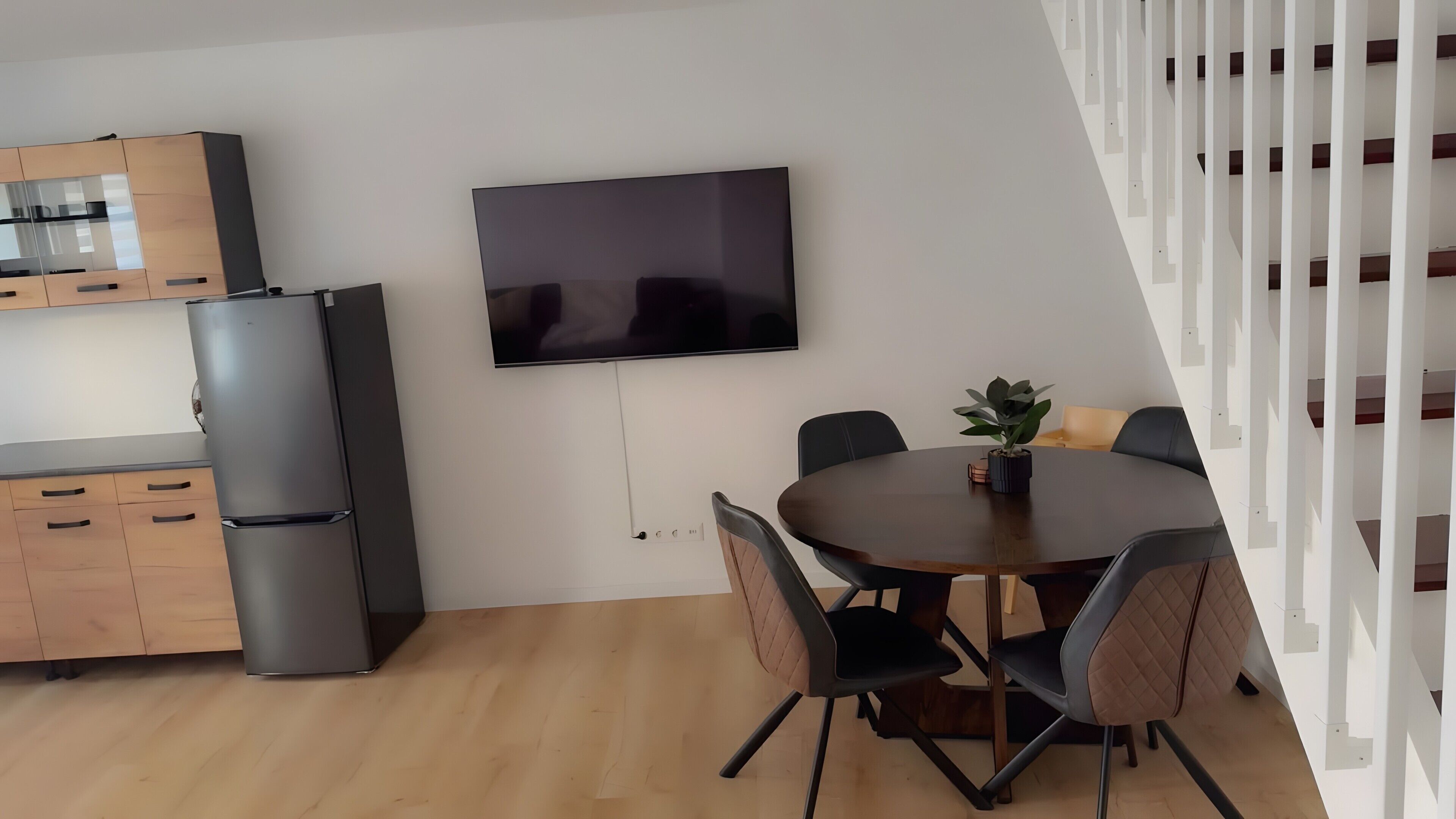 A modern kitchen with a built-in refrigerator, a dining area with a round table and chairs, and a flat-screen TV mounted on the wall.