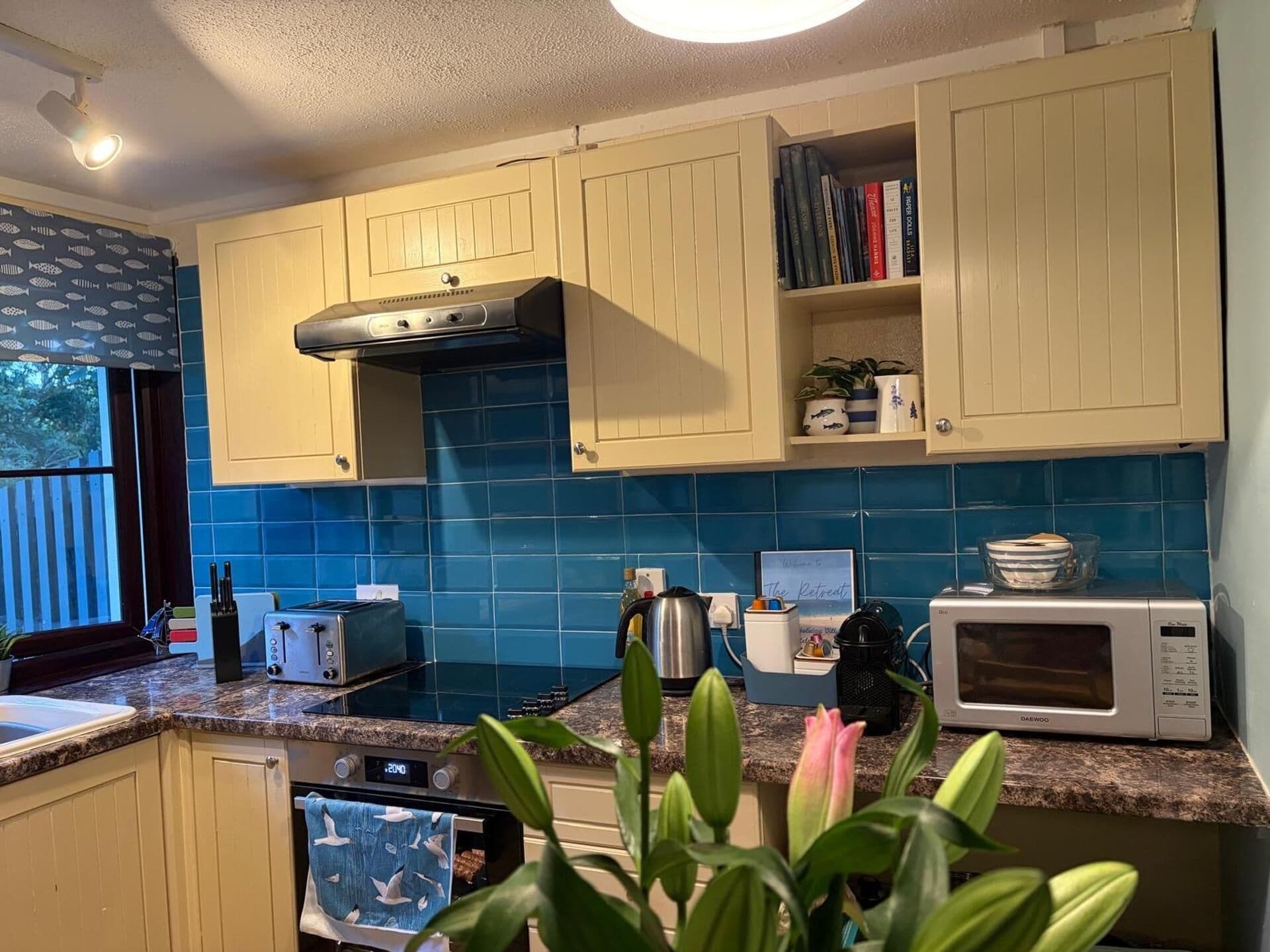 Una cocina con armarios de colores claros, salpicadero de azulejos azules, un tostador, un hervidor, un microondas y una ventana con vistas.