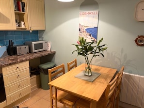 Una cocina con una mesa de madera, sillas, un microondas, un cubo de basura y un cartel en la pared.