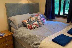 Una cama con ropa de cama a rayas y almohadas florales, una mesita de noche de madera con un libro, y una ventana con cortinas.