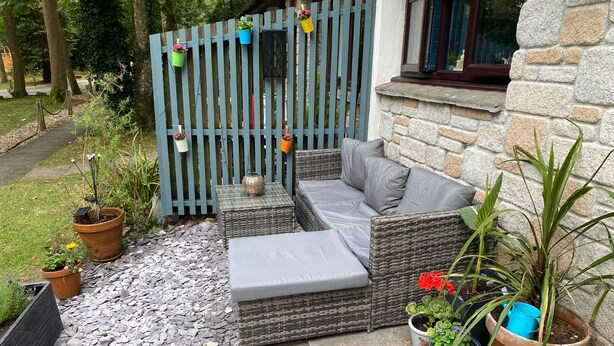 Eine Terrasse mit grauem Sofa, Korbmöbeln, Blumentöpfen und einem blauen Zaun.