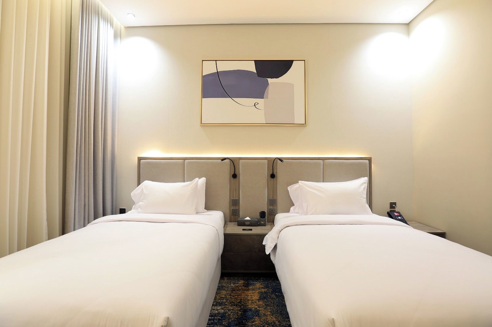 Een hotelkamer met twee bedden, een hoofdeinde met ingebouwde verlichting, een nachtkastje met een lamp en een schilderij boven het hoofdeinde.