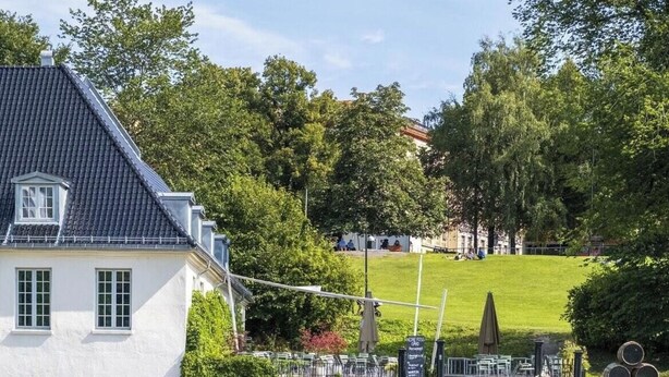 Un edificio blanco con techo oscuro, un área verde con valla y árboles al fondo.