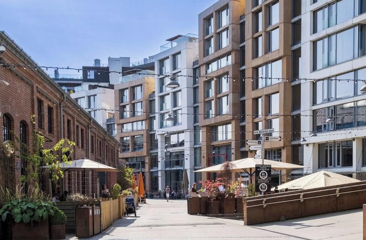 A modern urban street with multi-story buildings, outdoor seating areas, and a clear sky.